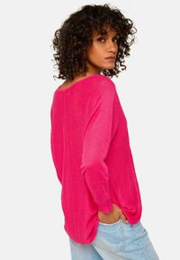 Helles pinkes Langarmpullover mit lockerer Passform, weitem Ausschnitt und strukturiertem Stoff. Das Model trägt ihn in die hellblauen Jeans gesteckt.