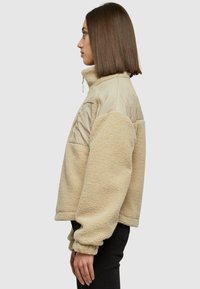 Osoba s ravnom smeđom kosom nosi bež flis jaknu s prošivenim panelima na ramenima, stoji u profilu na jednobojnoj pozadini.