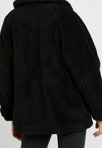 Czarny płaszcz z polaru o fakturowanym, puszystym wykończeniu, w oversize'owym kroju, z okrągłym kołnierzem, opadającymi ramionami oraz długością hemline sięgającą bioder.