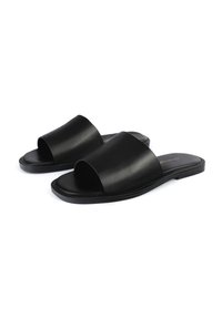 Des sandales noires à glissière en cuir lisse, présentant une large lanière avant et un design ouvert au niveau des orteils, avec une semelle en caoutchouc plate et un style minimaliste.