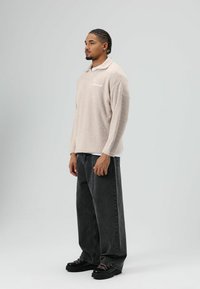 Beige fleece pullover met een kraag, voorzien van lange mouwen en een wit logo. Gecombineerd met loszittende donkergrijze jeans en zwarte schoenen.