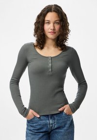 Giovane donna con capelli ricci che indossa una maglia grigia aderente a maniche lunghe con bottoni e jeans blu, con le mani nelle tasche, rivolta in avanti.