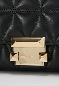 Černá prošívaná kožená kabelka se zapínáním zlaté barvy s hroty, raženým logem a zdobeným prošíváním.