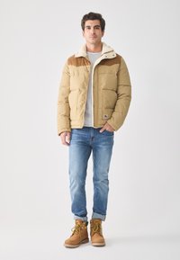 Hombre de pie con chaqueta acolchada color beige y cuello de borrego, camiseta gris claro, jeans azules y botas de trabajo beige, sobre fondo blanco liso.
