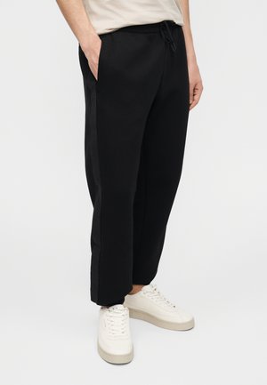 Man met zwarte broek met trekkoord en zijstrepen en witte sneakers, staand met handen in de zakken tegen een effen achtergrond.