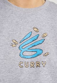 Sweat-shirt gris avec un design graphique bleu représentant un 'C' stylisé et des illustrations de cookies, centré au-dessus du mot "CURRY".