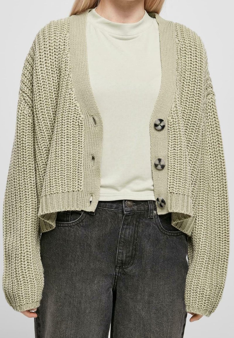 Femme portant un cardigan en grosse maille beige oversize avec de gros boutons, par-dessus un col roulé crème clair et un jean foncé délavé.
