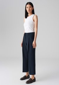 Un débardeur blanc ajusté, un pantalon taille haute marine foncé avec des plis, et des mocassins marron. Tissu lisse, coupe sur mesure, longueur de jambe raccourcie.