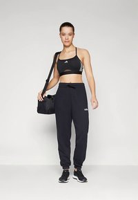 Jovem mulher em top desportivo preto, calças de treino Adidas pretas, ténis pretos, com uma mala de ginásio preta ao ombro, de pé contra um fundo simples.