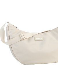 Beige Stoff-Schultertasche mit einer geschwungenen Form, verstellbarem Tragegurt und einem Logo-Patch. Glatte Oberfläche mit einem Reißverschluss oben.