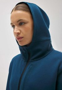 Sudadera de color verde oscuro con un alto cuello y capucha integrada. Presenta una cremallera negra y una textura de tela suave. Sin patrones ni acentos visibles.