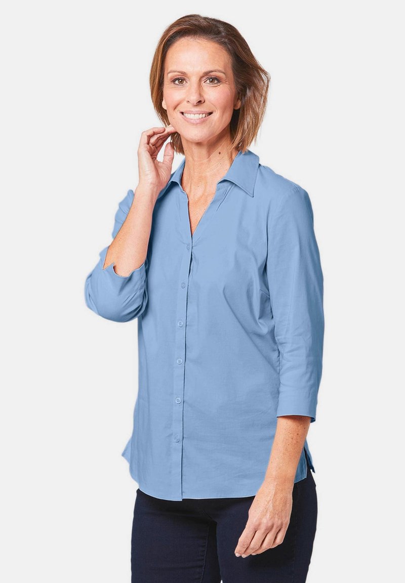 Camicia azzurra chiaro con colletto e maniche tre quarti. Il tessuto appare liscio, con una vestibilità comoda e spacco laterale per facilitare i movimenti.