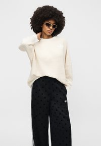 Γυναίκα με μεγάλο αφρό μαλλιών που φοράει μπεζ πλεκτό πουλόβερ, μαύρο παντελόνι με άσπρες πουά και γυαλιά ηλίου cat-eye, ποζάροντας με το ένα χέρι πίσω από τον αυχένα.