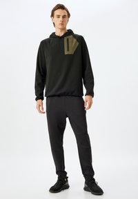 Sudadera negra con capucha y un bolsillo de acento verde, combinada con pantalones gris oscuro de corte ajustado y tejido texturizado, llevados con zapatillas negras.