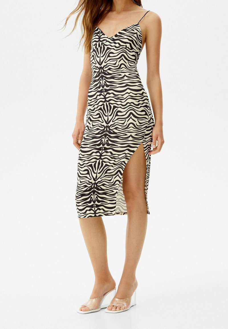Robe midi imprimée zèbre en crème et noir avec des bretelles fines, un décolleté en V et une fente sur le côté. Le mannequin porte des sandales à talons blocs transparents.