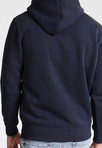Tmavě modrá bavlněná mikina s kapucí na stahovací šňůrku, žebrovanými manžetami a lemem, s hladkou texturou bez viditelných log nebo ozdob.