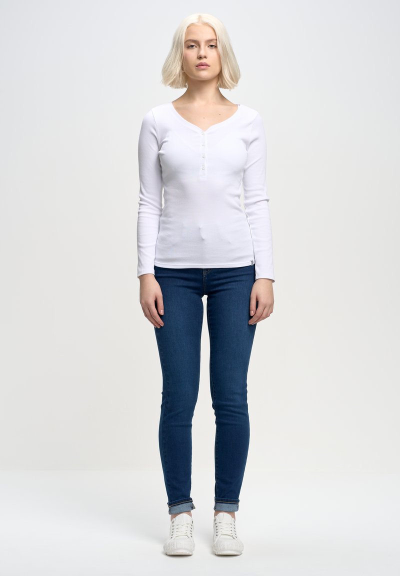 Witte lange mouw top met een V-hals en knoopaccenten, gecombineerd met donkerblauwe skinny jeans en witte sneakers, tegen een effen achtergrond.