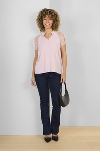 Blusa rosa chiaro con maniche in pizzo, scollatura a V, abbinata a jeans blu scuro e scarpe nere con punta, che tiene una borsa nera.
