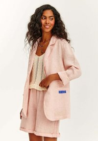 Světle růžový lněný blejzr s klopovým límcem, našitými kapsami a modrou nášivkou; nošený přes krémový pletený top a odpovídající šortky.