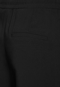 Gros plan sur un pantalon en tissu noir présentant une poche arrière fendue horizontalement avec des coutures visibles et un détail de ceinture élastique.