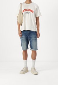 Tričko v barvě off-white s červeným logem, denimové šortky s ohrnutými manžetami, bílé kotníkové ponožky a krémové tenisky; béžová taška přes rameno.