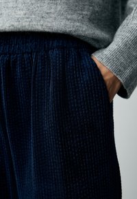 Pantaloni di velluto a coste blu navy con superficie testurizzata, dotati di elastico in vita e tasche laterali. Maglione lavorato a maglia grigio parzialmente visibile sopra.