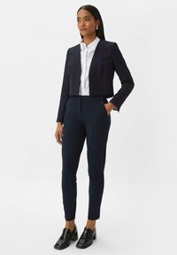 Marineblauwe cropped blazer met gestructureerde schouders, gecombineerd met slim-fit marineblauwe pantalons en glanzende zwarte loafers, die de strakke lijnen en de op maat gemaakte pasvorm benadrukken.