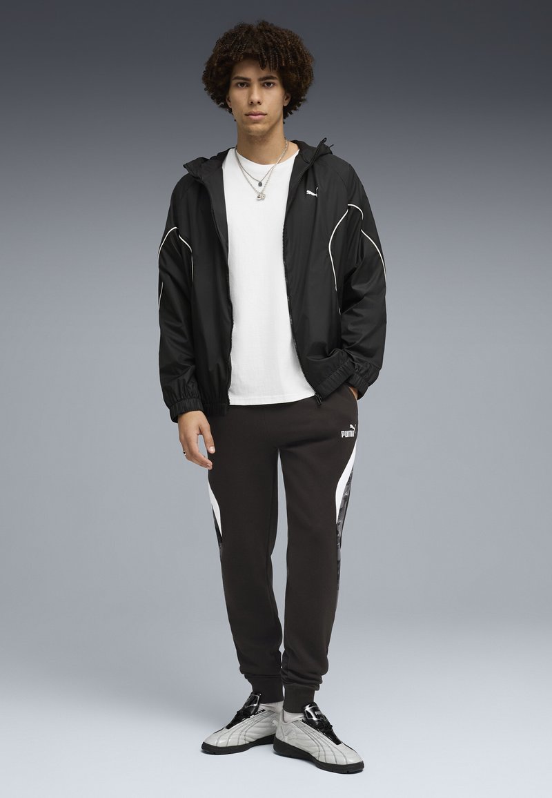 Junger Mann steht vor grauem Hintergrund, trägt eine schwarze Puma-Windjacke, ein weißes T-Shirt, schwarze Puma-Sweatpants mit weißen Streifen und weiße Turnschuhe.