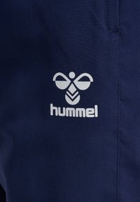 Weißes Hummel-Markenlogo, das einer stilisierten Biene auf marineblauem Stoff ähnelt, wahrscheinlich Teil eines Kleidungsstücks mit sichtbaren Nahtnähten.