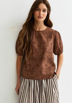 Jonge vrouw die een bruine geborduurde blouse met korte mouwen en een gestreepte geplooide rok draagt, staand tegen een witte muur met haar hand in de zak.