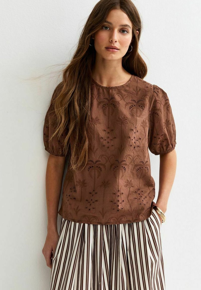 Young woman wearing a brown embroidered short-sleeve blouse and striped pleated skirt, standing against a white wall with hand in pocket.