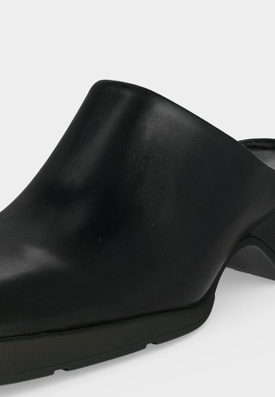 Μαύρο mule slip-on παπούτσι με λείο δερμάτινο πάνω μέρος και χοντρό χαμηλό τακούνι, απεικονίζεται σε απλό ανοιχτό φόντο.