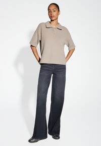 Pull en laine beige mélangé avec des manches courtes et des détails de boutons, associé à un jean flare bleu foncé et des chaussures noires.