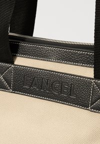 Geantă de pânză cu margini din piele neagră și logo embosat. Prezintă mânere negre și o broderie în contrast pe un fundal bej.