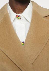 Dunkelhäutige Person in einem weißen Hemd mit einem bunten geometrischen Kragen-Pin und einem kamelfarbenen Wollmantel.