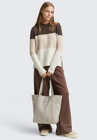 Gehaakte kleurblokkensweater in bruin, beige en crème; losse pasvorm met ribbelrand. Grijze handtas; brede bruine broeken. Crème schoenen.