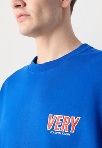 Sweatshirt azul de algodão com gola redonda, apresentando o texto bordado em vermelho "VERY" e "CALVIN KLEIN" do lado esquerdo. Textura suave.