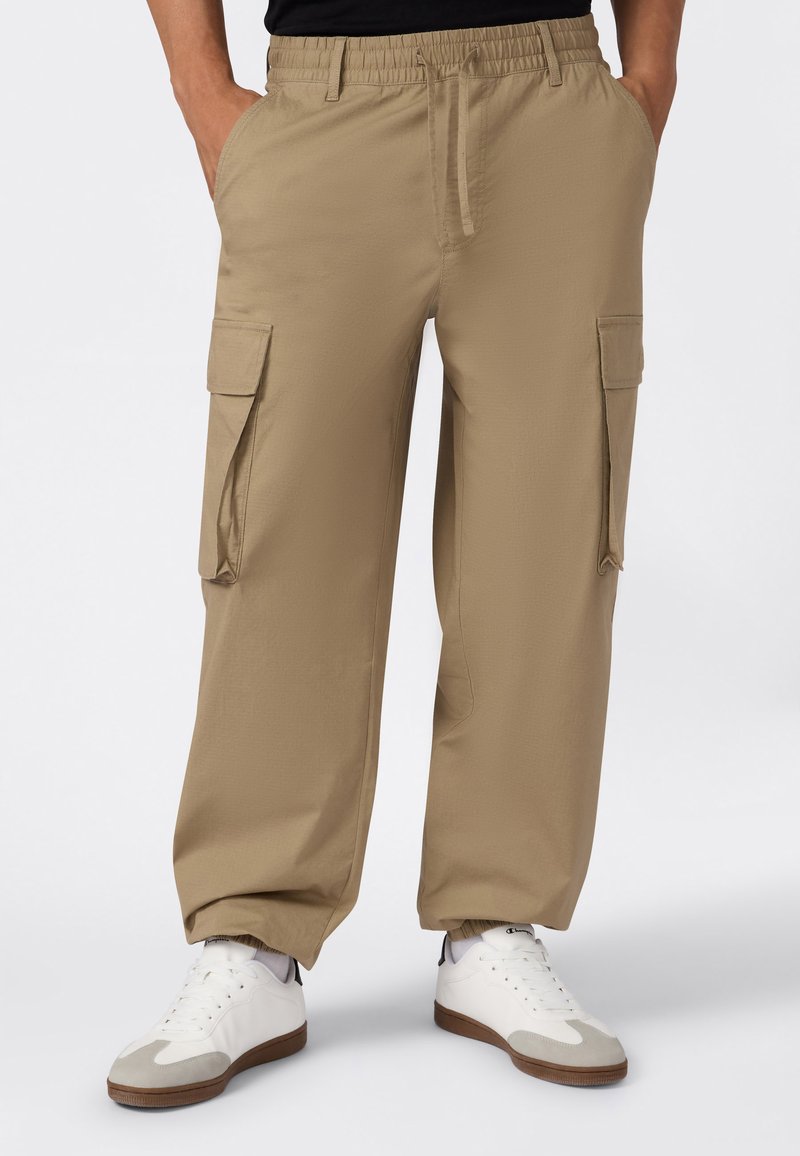 Onderlichaam gekleed in beige cargobroek met zijzakken, elastische tailleband en witte sneakers met grijze en bruine accenten.
