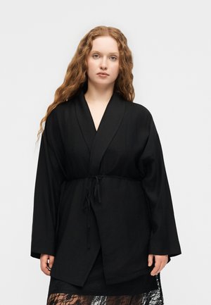 Γυναίκα με μακριά σγουρά κόκκινα μαλλιά, που φοράει μαύρο χαλαρό σακάκι τύπου wrap δεμένο στη μέση και μαύρη φούστα από δαντέλα, στέκεται μπροστά σε απλό φόντο.