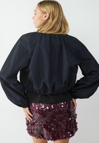 Schwarze Bomberjacke mit voluminösen Ärmeln und elastischem Bund, kombiniert mit einem bordeauxroten, mit Pailletten besetzten Minirock, der reflektierende, kreisförmige Verzierungen präsentiert.