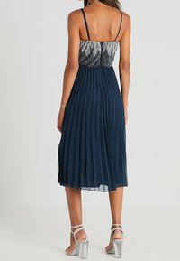 Námořnické plisované šaty s perlovým stříbrným korzetem, tenkými ramínky a délkou do poloviny lýtek. Vzhled doplňují stříbrné sandály na podpatku.