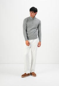 Mann in einem grauen Halbzip-Pullover, weißen Hosen und braunen Slippern, der vor einem schlichten weißen Hintergrund steht.