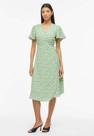 Femme portant une robe portefeuille fleurie vert clair avec de courtes manches volantées et des mules à talons noires à bout ouvert, debout devant un fond uni.