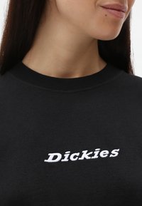 Svart hettegenser med hvit brodert "Dickies" tekst, med rund hals og en jevn tekstur. Nærbilde av det øvre plagget.