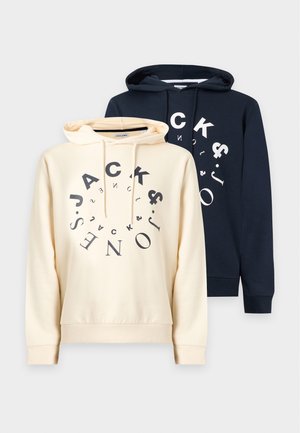 Mann trägt schwarzen Hoodie mit rundem "Jack & Jones"-Logo, lockere blaue Jeans und weiße Sneakers, steht vor weißem Hintergrund.