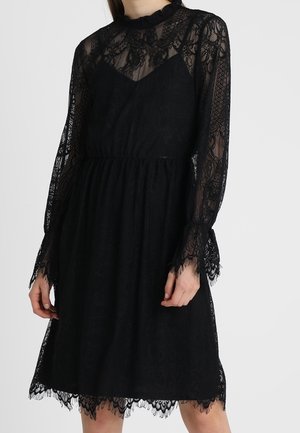 Frau trägt ein knielanges schwarzes Spitzenkleid mit langen transparenten Spitzenärmeln und hohem Ausschnitt.