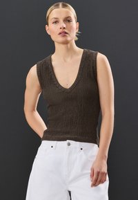 Braunes ärmelloses Oberteil mit einer strukturierten Strickmuster und V-Ausschnitt, kombiniert mit weißen hoch taillierten Hosen mit Knopfdetails.