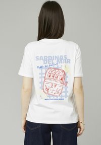 T-shirt en coton blanc avec un graphique coloré de sardines en conserve, accompagné de texte en bleu et rouge, le tout accentué par un motif de grille.
