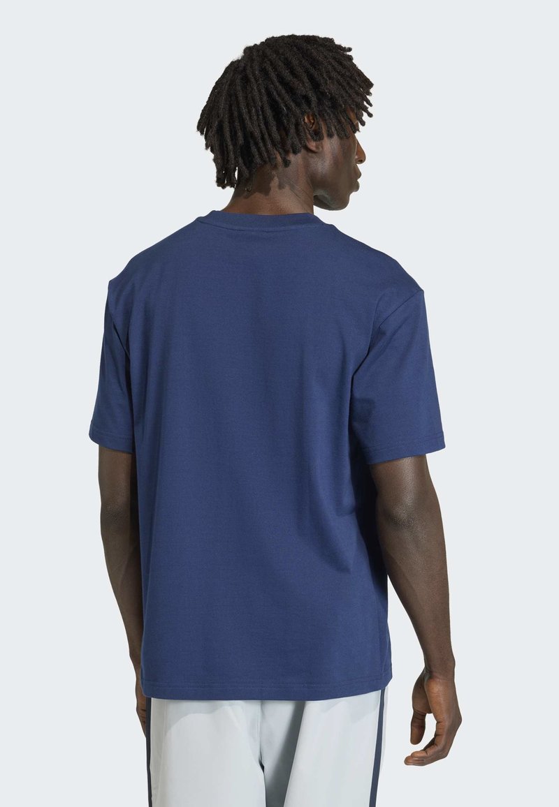 Uomo con pelle scura e dreadlocks corti che indossa una maglietta semplice blu navy a maniche corte e pantaloni grigio chiaro, in piedi di spalle.
