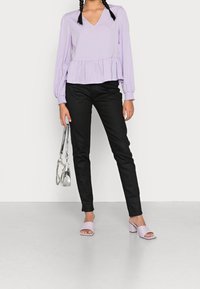 Atasan peplum lavender dengan lengan panjang, dipadukan dengan celana hitam slim-fit dan mule bertumit blok lavender, sambil memegang tas tangan perak.
